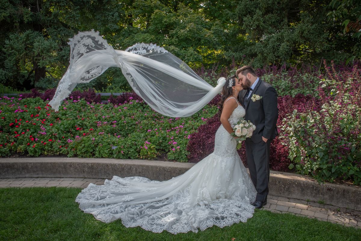 A bride and groom kiss in a garden with a billowing veil and vibrant flowers under dappled sunlight.