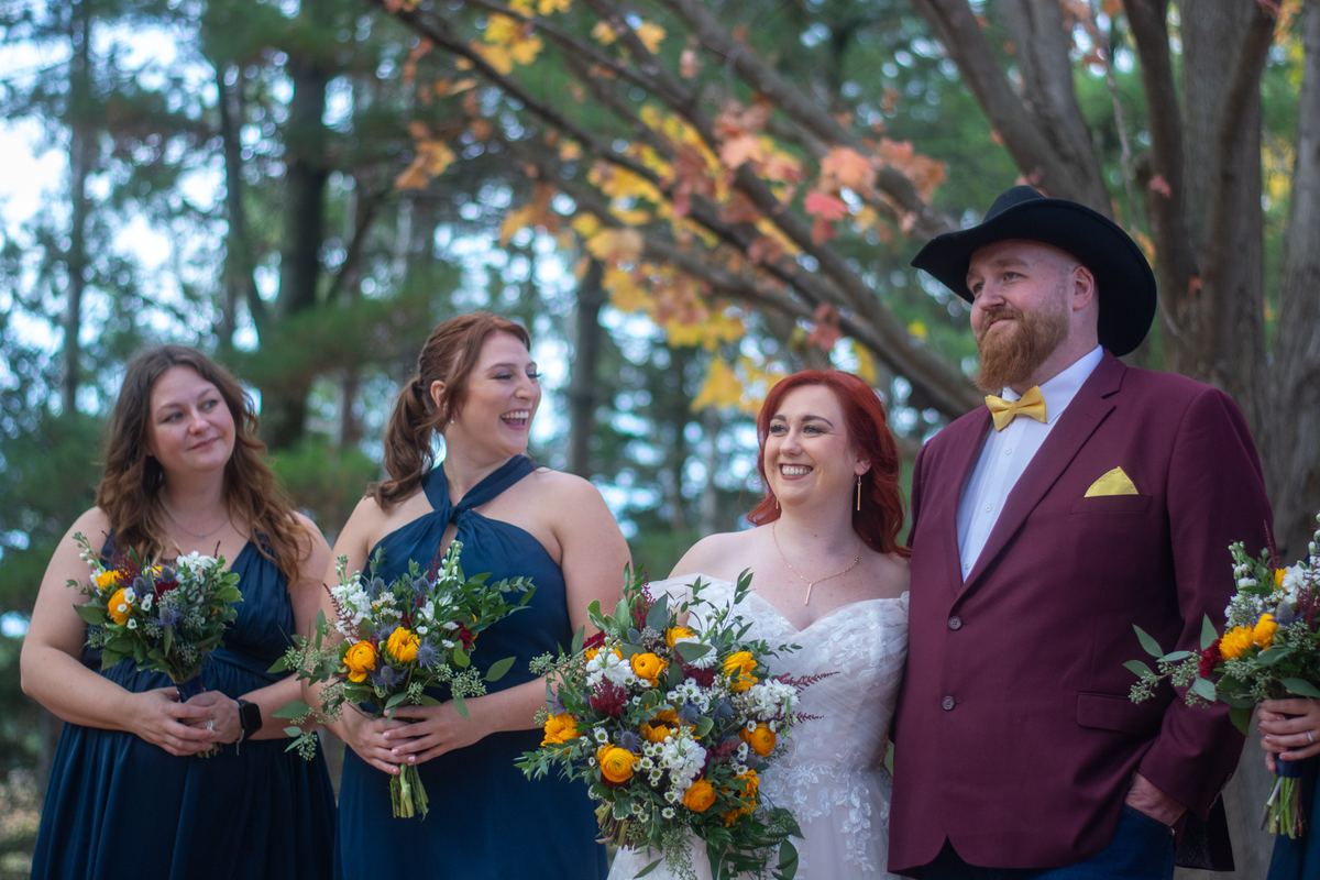 A wedding party poses outdoors under autumn trees, smiling and laughing with colorful flower bouquets.