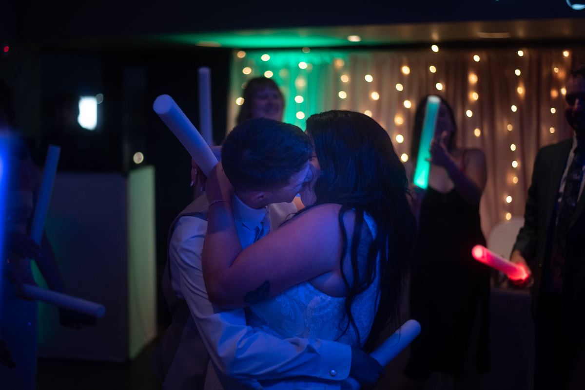 A couple kisses at a celebration with guests holding glowing sticks and string lights in the background.