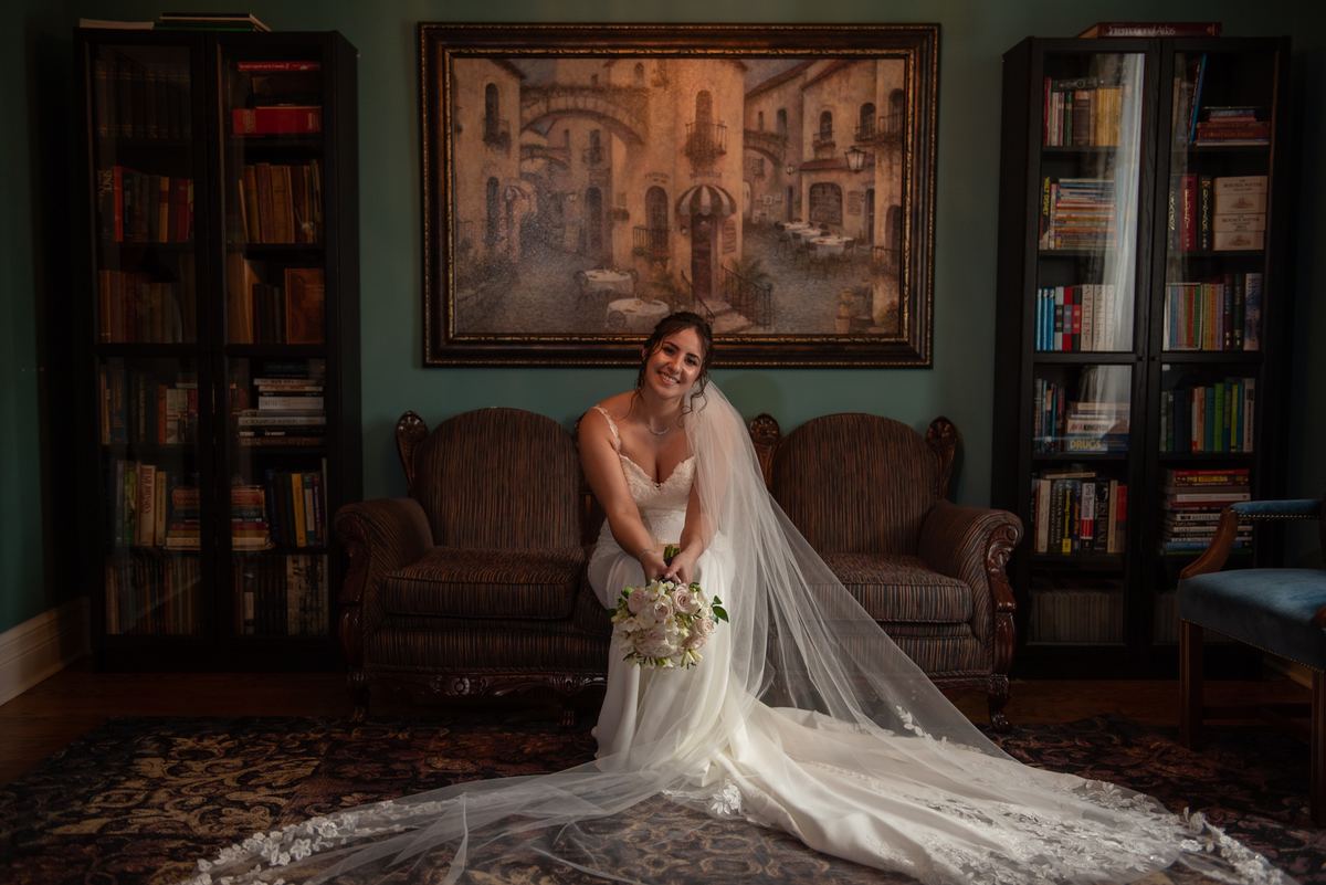 A smiling bride sits on a vintage sofa in an ornate room, holding her bouquet and surrounded by books.