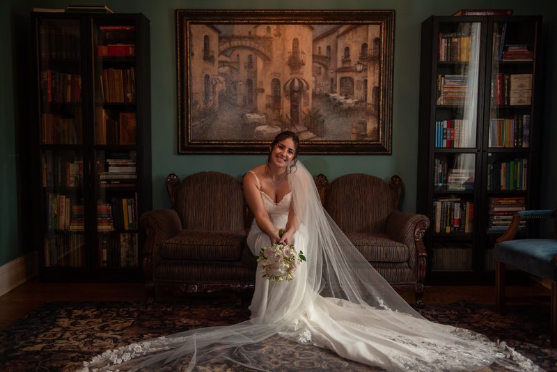 A smiling bride sits on a vintage sofa in an ornate room, holding her bouquet and surrounded by books.