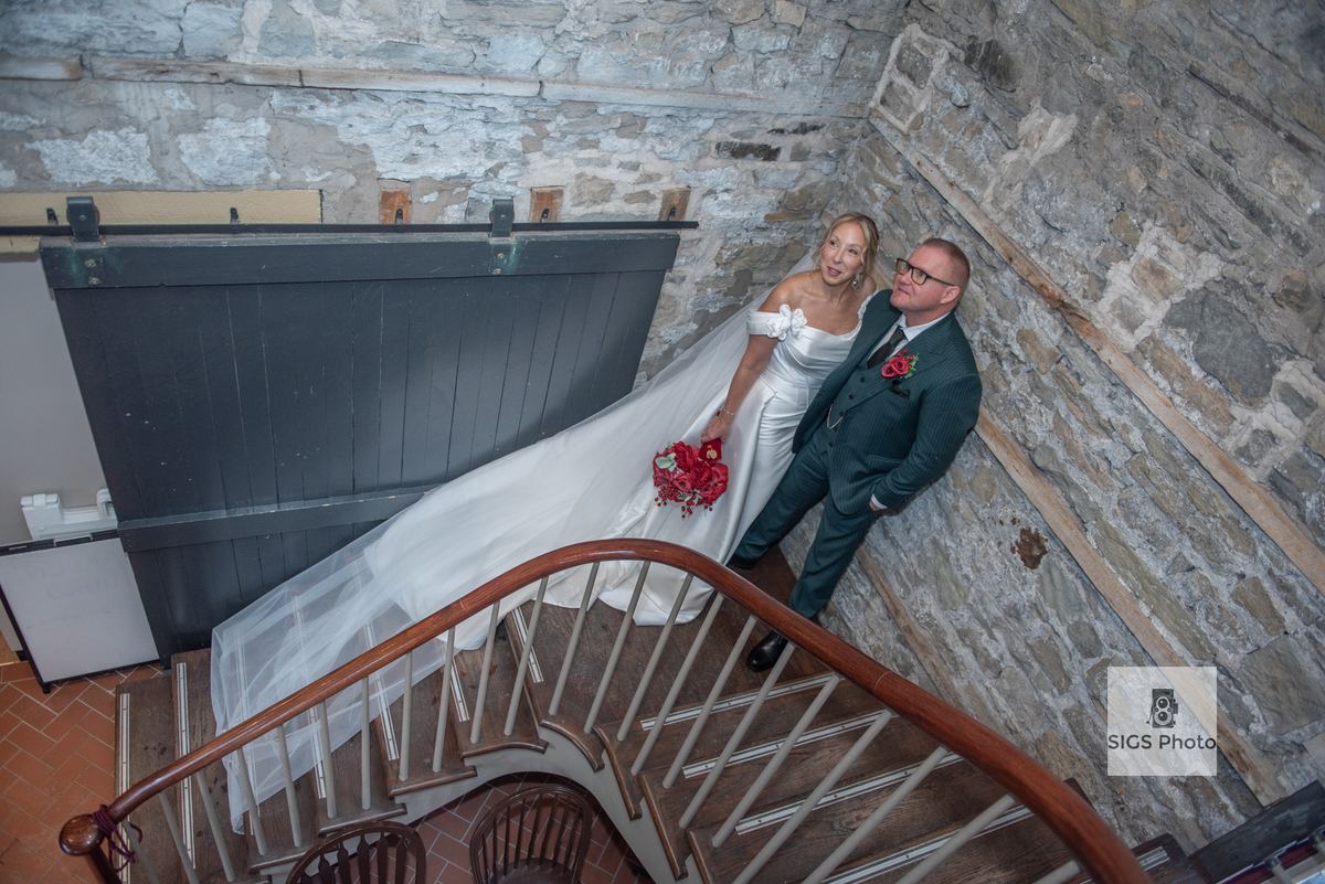 Bride in white gown and groom in dark suit smile warmly on wooden staircase inside rustic stone building.
