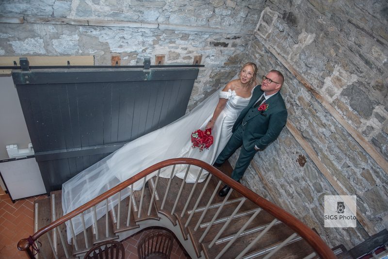 Bride in white gown and groom in dark suit smile warmly on wooden staircase inside rustic stone building.