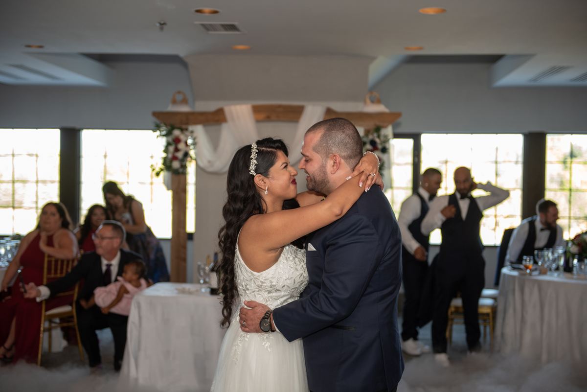 Bride and groom dance closely, surrounded by guests at a reception with natural light streaming through windows.