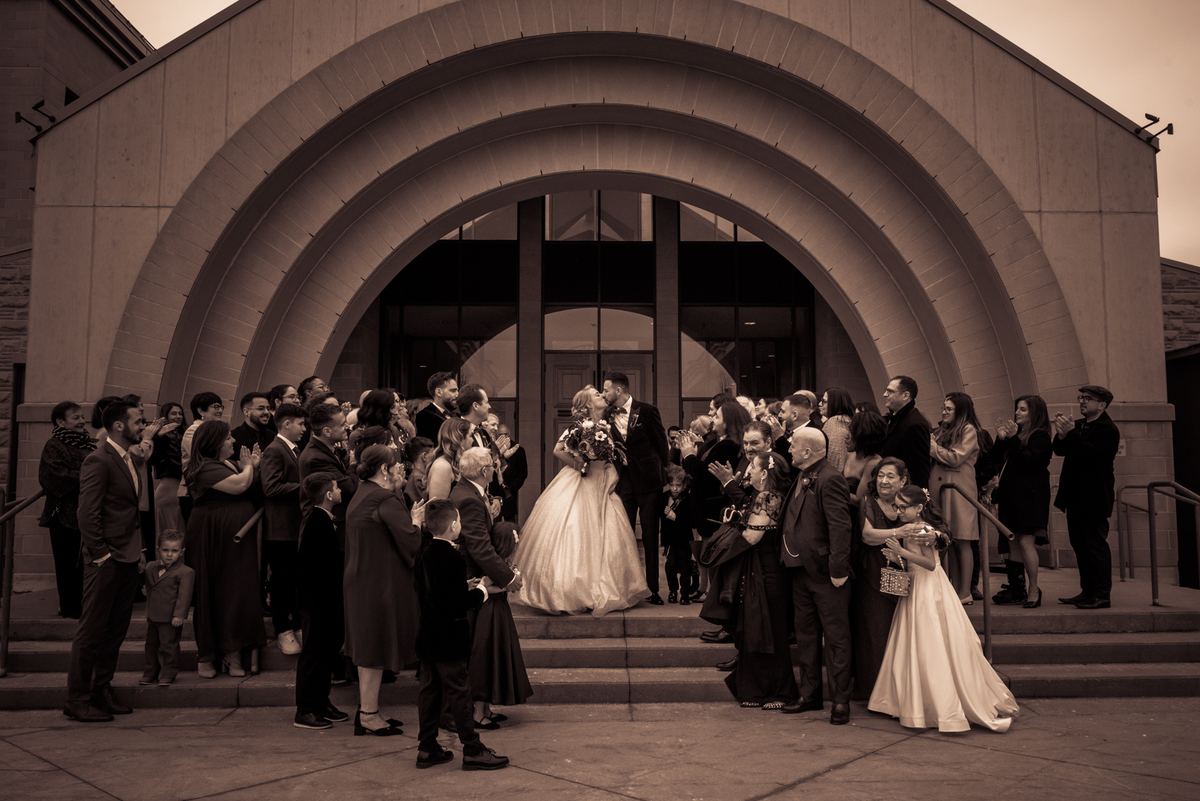A newlywed couple kisses passionately on steps, surrounded by smiling guests applauding in warm, sepia-toned light.