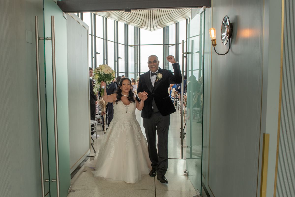 Bride and groom walk down glass-enclosed aisle toward camera, groom raises fist, bride smiles with bouquet.