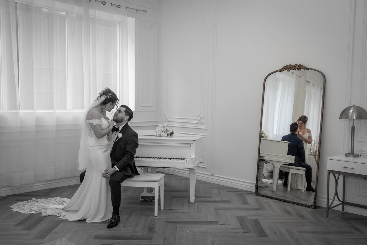 A bride and groom share a tender moment beside a grand piano in an elegant room, reflected in a large mirror.