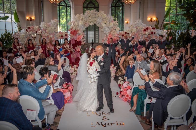 Maria and Mike share a kiss at the altar beneath a lavish floral arch, surrounded by cheering guests.