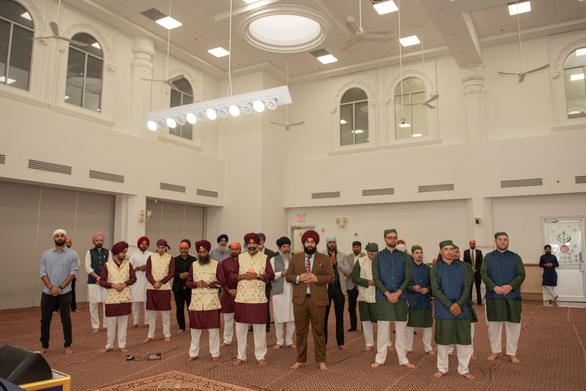 Men in traditional South Asian attire stand in prayerful gestures within a grand hall with arched windows.