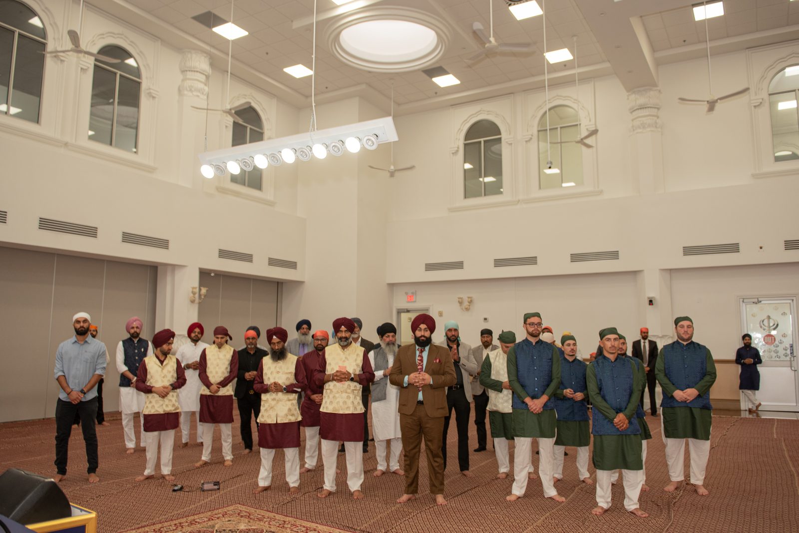 Men in traditional South Asian attire stand in prayerful gestures within a grand hall with arched windows.