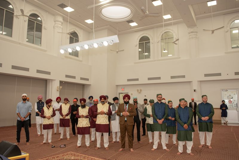 Men in traditional South Asian attire stand in prayerful gestures within a grand hall with arched windows.