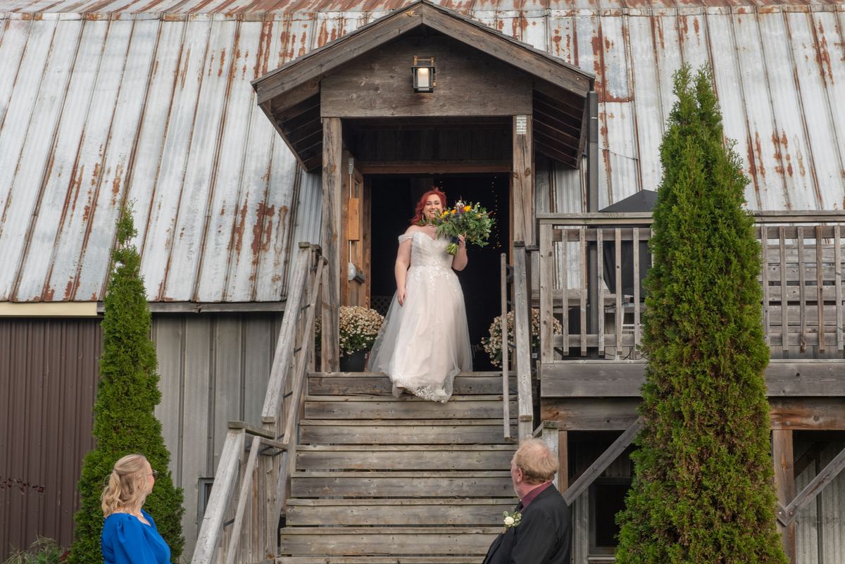 A red-haired bride in a white gown descends wooden steps from an old barn, approaching two guests on a softly lit path.