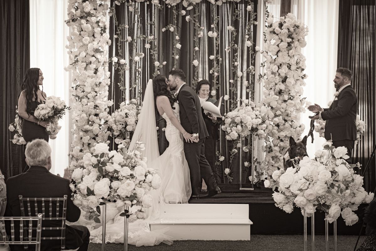 A bride and groom kiss at their wedding altar, surrounded by flowers and drapes, with guests observing.
