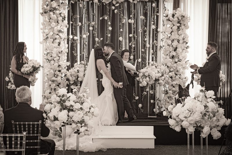 A bride and groom kiss at their wedding altar, surrounded by flowers and drapes, with guests observing.