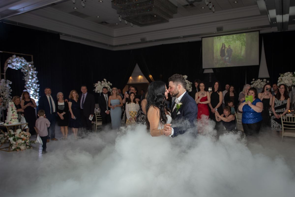 A bride and groom dance intimately as guests watch from the sidelines, surrounded by white smoke and floral decorations.