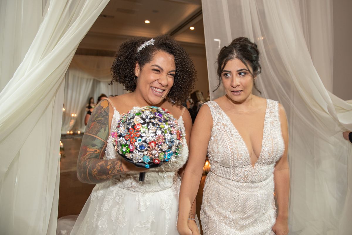 Two brides smiling and laughing under a draped archway, one holding a sparkling bouquet, in warm ambient lighting.
