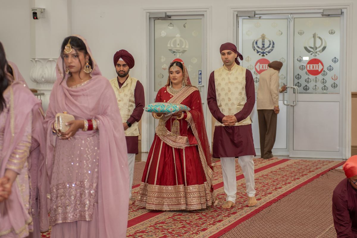 A bride in red and gold holds a pillow with the groom, surrounded by attendants in traditional attire within a Sikh gurdwara.