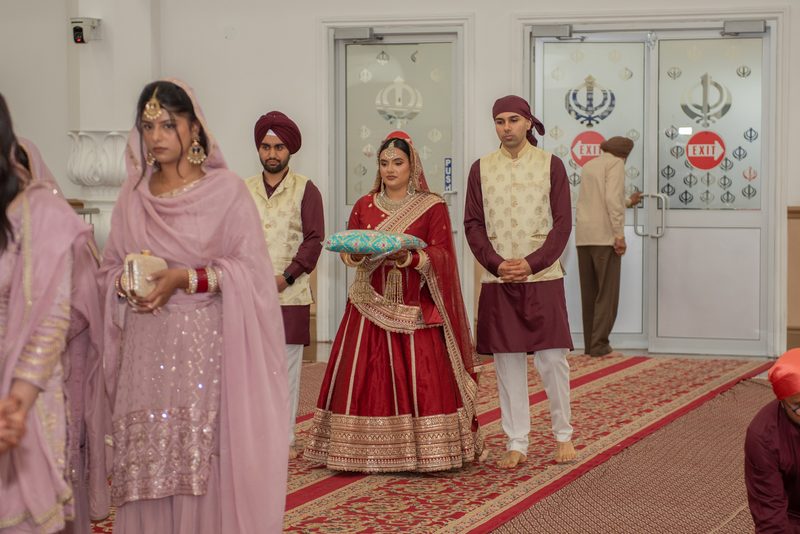A bride in red and gold holds a pillow with the groom, surrounded by attendants in traditional attire within a Sikh gurdwara.