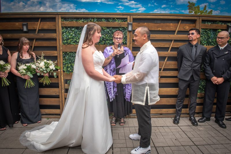A bride and groom hold hands during an outdoor wedding ceremony under a blue sky, with guests in formal attire nearby.