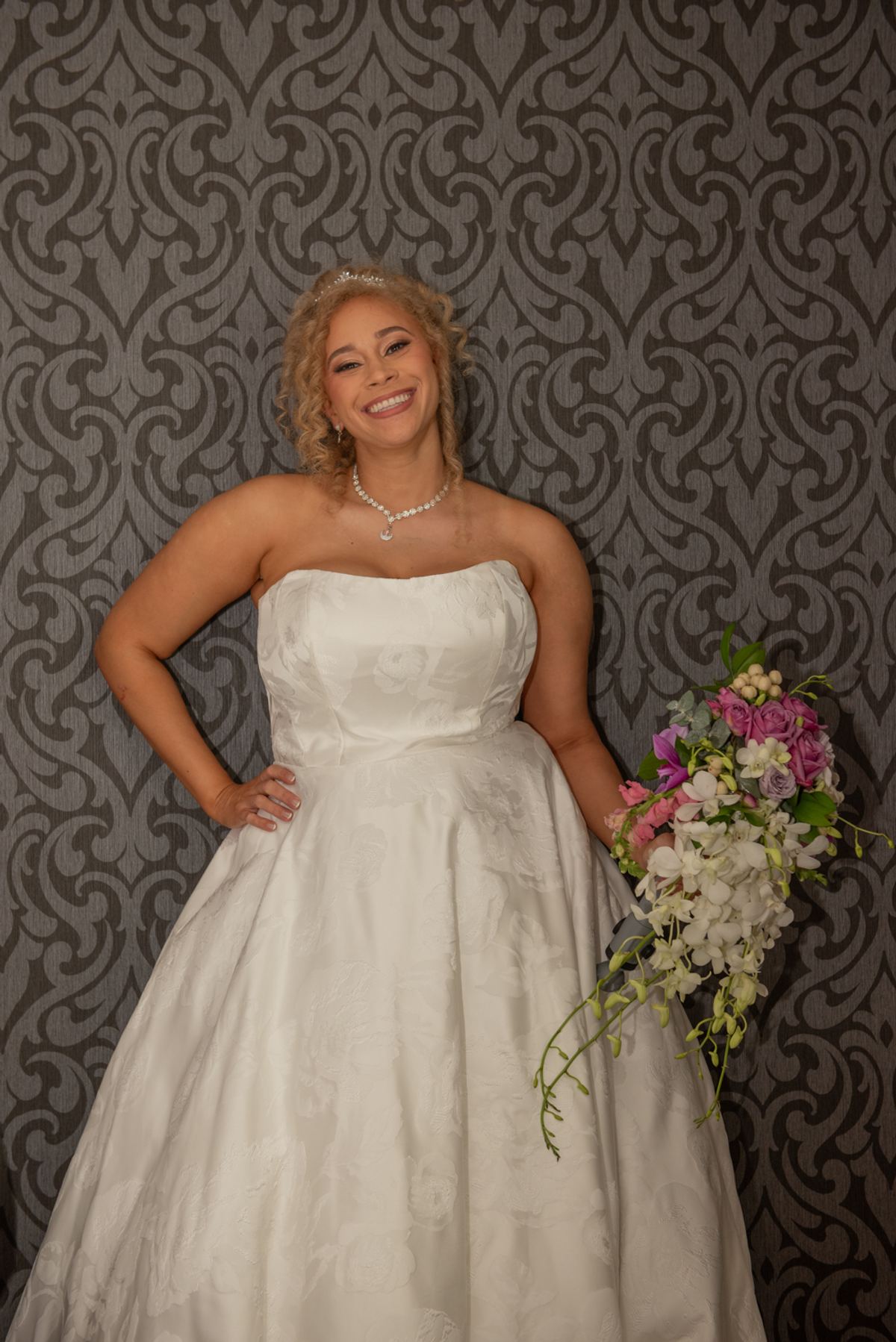 A smiling bride in a white wedding gown with a colorful bouquet stands confidently against ornate wallpaper.