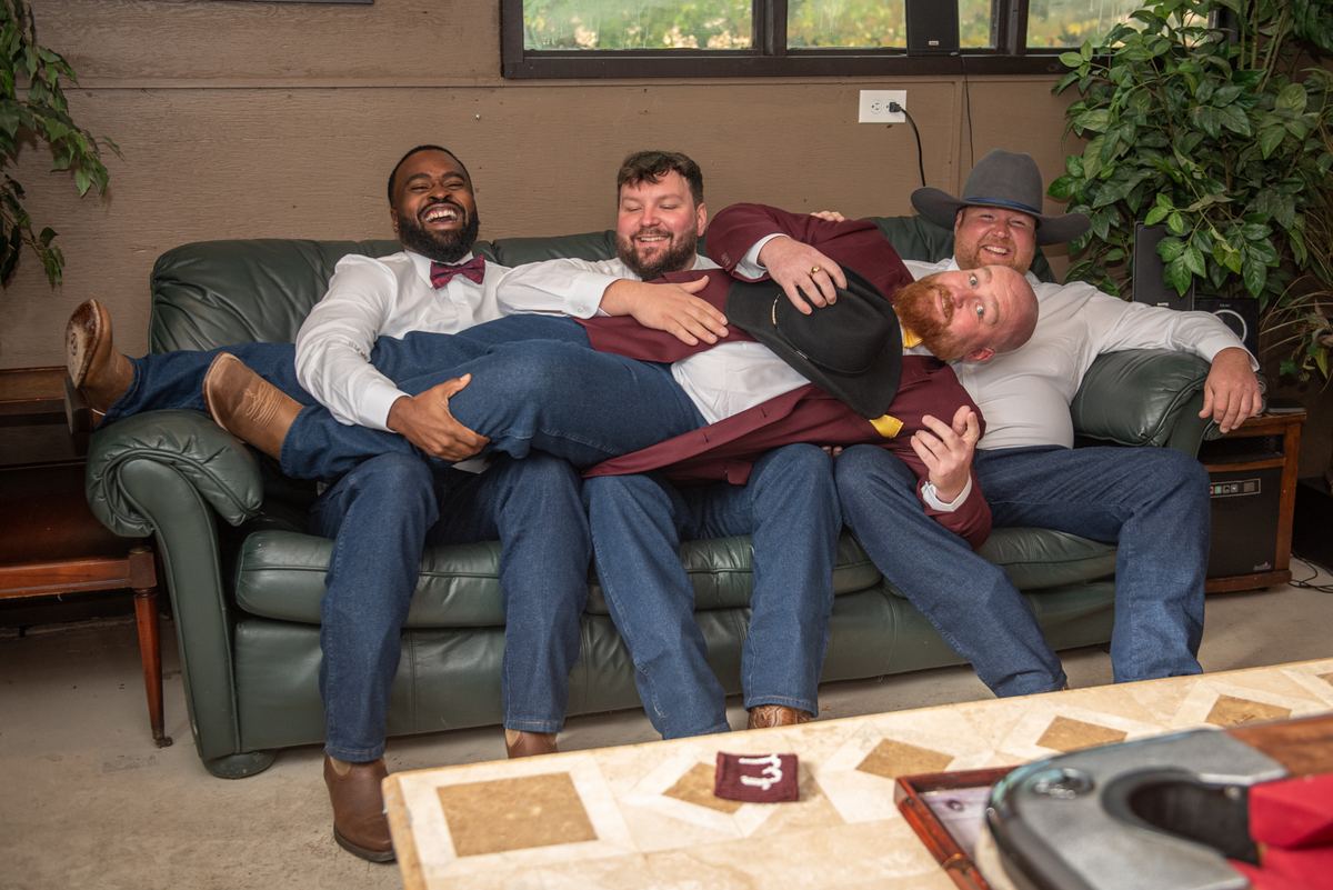 Four men in semi-formal attire, including bow ties and cowboy hats, laugh and embrace on a green leather couch.