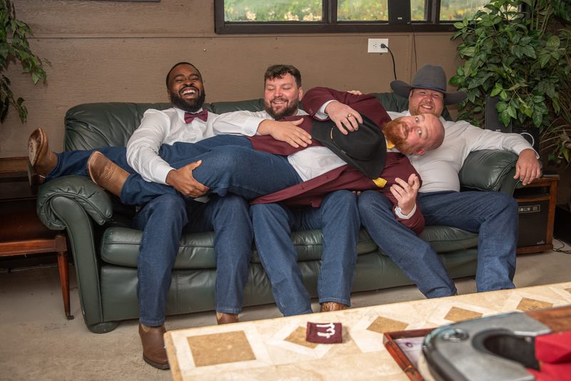 Four men in semi-formal attire, including bow ties and cowboy hats, laugh and embrace on a green leather couch.