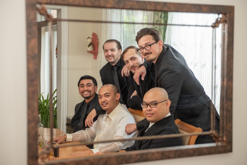 Six formally dressed men gather around a table, smiling and posing in front of an ornate mirror in a warmly lit room.