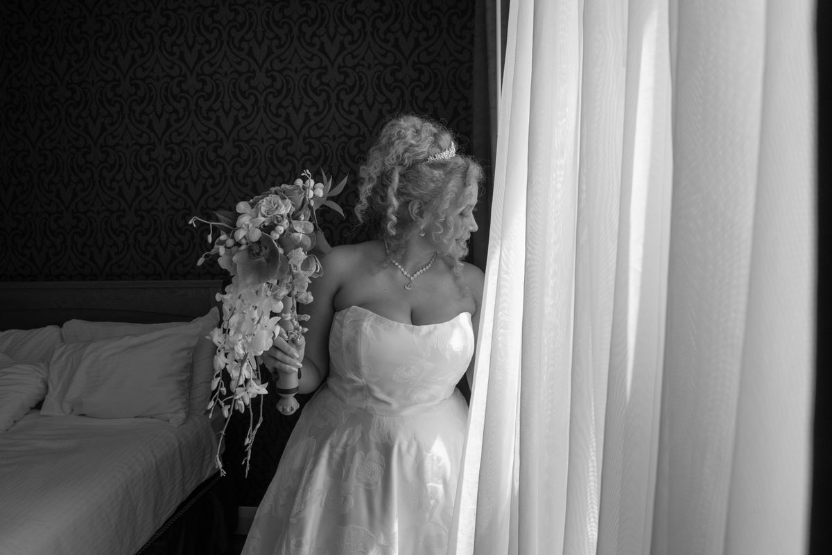 Bride in black and white stands by window, gazing through sheer curtains with cascading bouquet, illuminated by soft light.