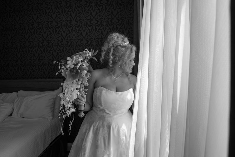 Bride in black and white stands by window, gazing through sheer curtains with cascading bouquet, illuminated by soft light.