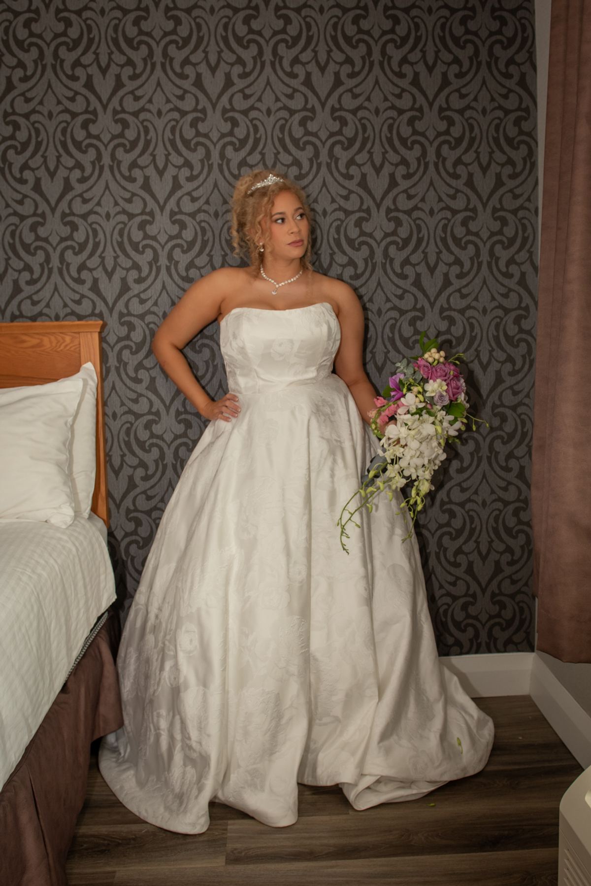A bride in a white gown holds pink and white flowers near an ornate wooden bed, standing alone in the room.