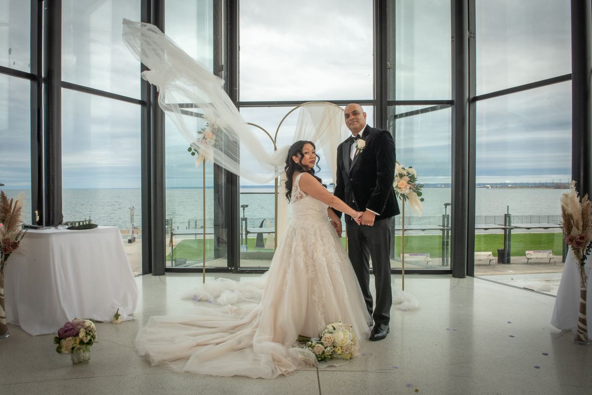 A bride and groom stand in a modern glass-walled space with water views, her veil billowing in the wind.