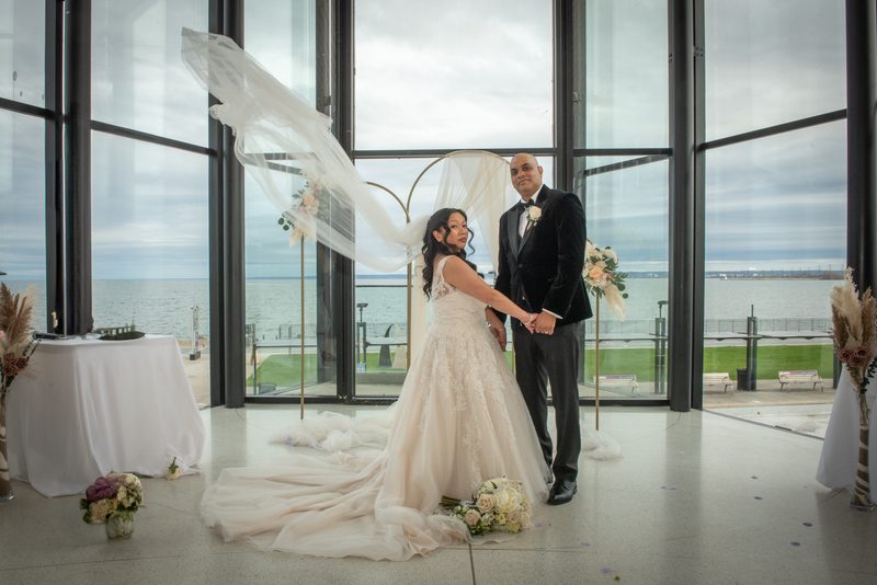 A bride and groom stand in a modern glass-walled space with water views, her veil billowing in the wind.