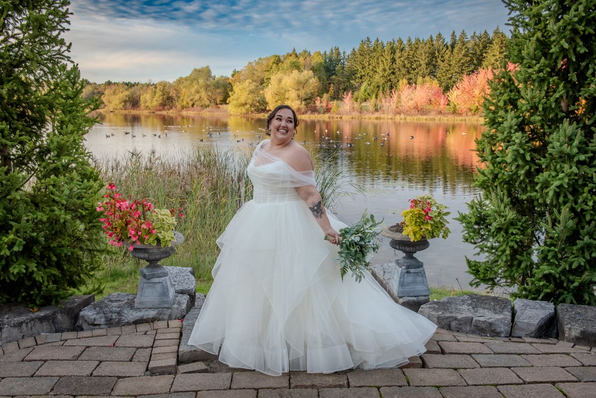 A smiling bride in a white gown stands on a stone patio by a lake, surrounded by trees and autumn leaves.