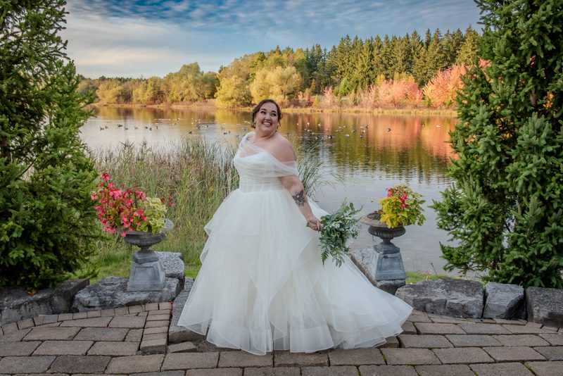 A smiling bride in a white gown stands on a stone patio by a lake, surrounded by trees and autumn leaves.