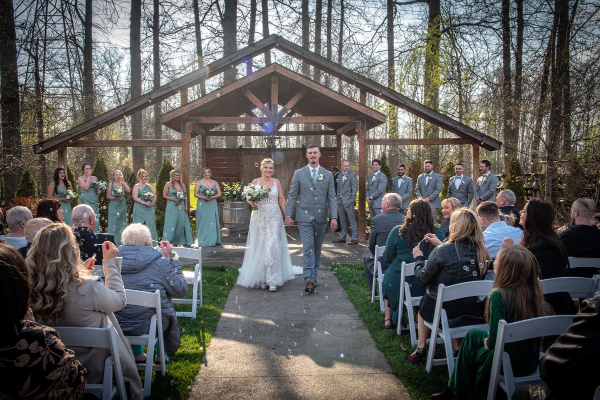 A bride and groom walk down the aisle under a wooden canopy, guests applauding as confetti floats in sunlight.