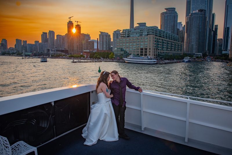A bride and groom stand on a ferry deck, gazing at each other as the sun sets behind Toronto's skyline.
