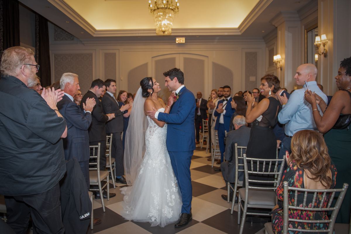 A bride and groom embrace in a ballroom as guests applaud, surrounded by formal attire-clad attendees.