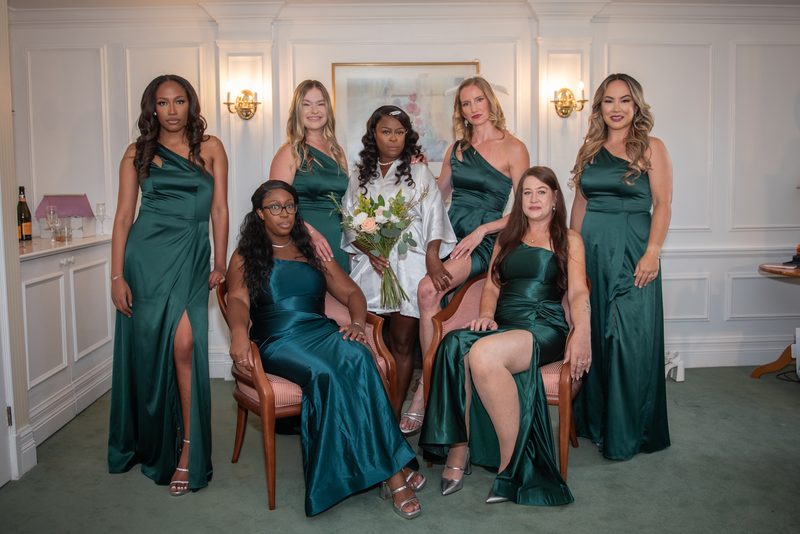 A bride in a white gown sits with her bridesmaids in emerald green dresses, illuminated by warm sconces.
