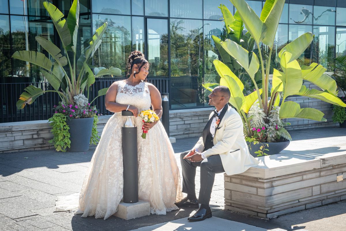 Bride in lace gown and groom in cream tuxedo stand beside stone bench in sunlit courtyard with tropical plants.