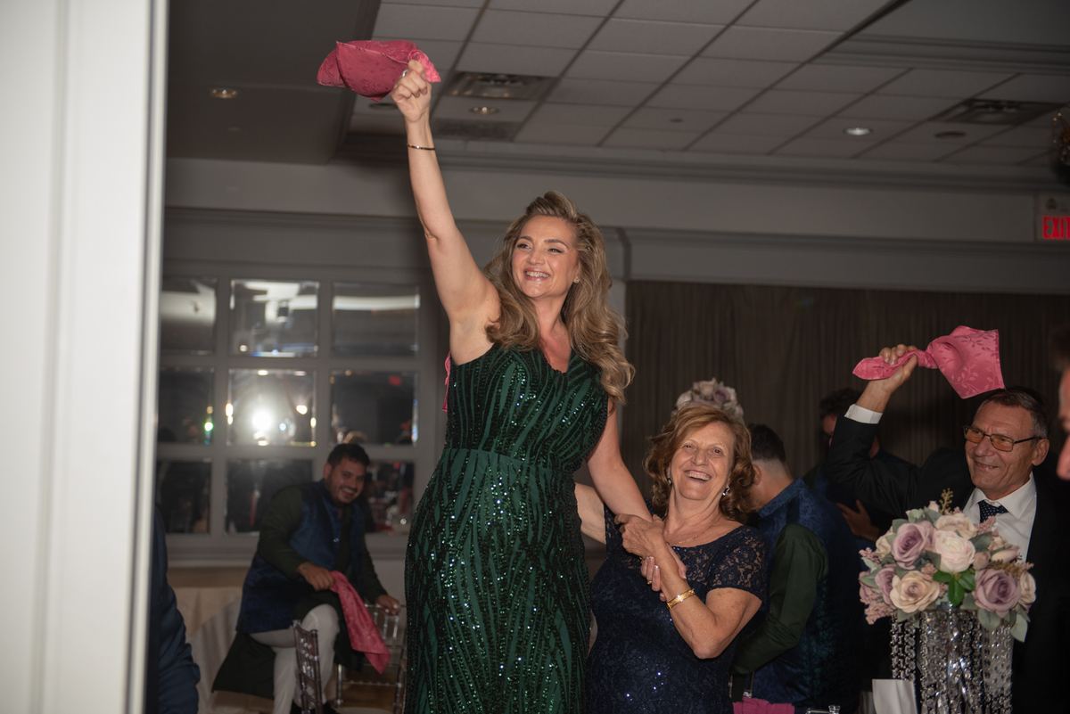 A joyful woman in a green sequined gown raises a pink cloth as guests, including a family member and a man in a suit, laugh.