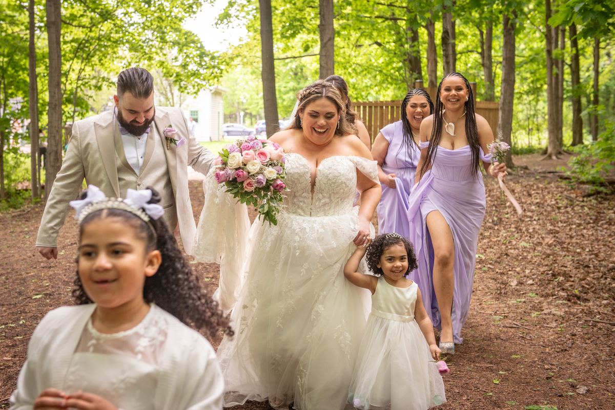 A bride laughs while holding a flower girl's hand, walking with her bridal party along a sunlit wooded path.