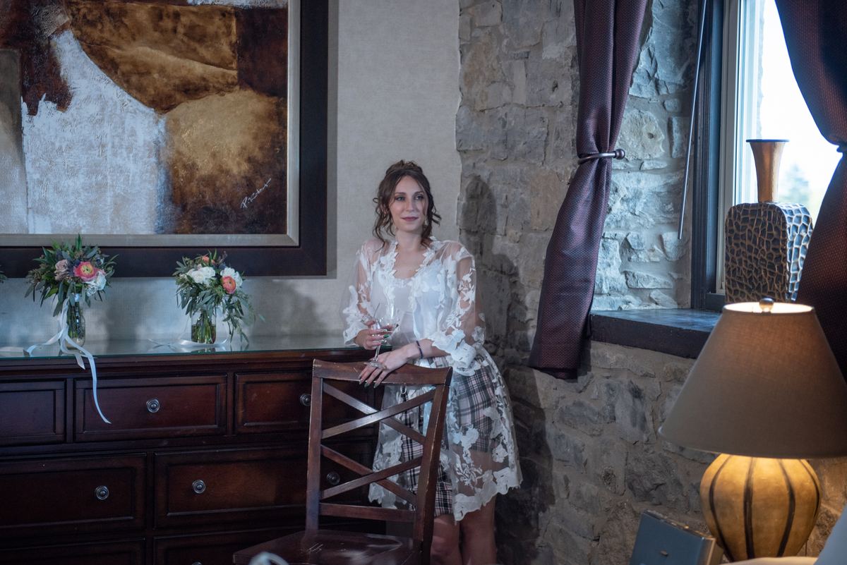 A woman in a white lace dress stands by a wooden chair in a rustic room with stone walls and an abstract painting.
