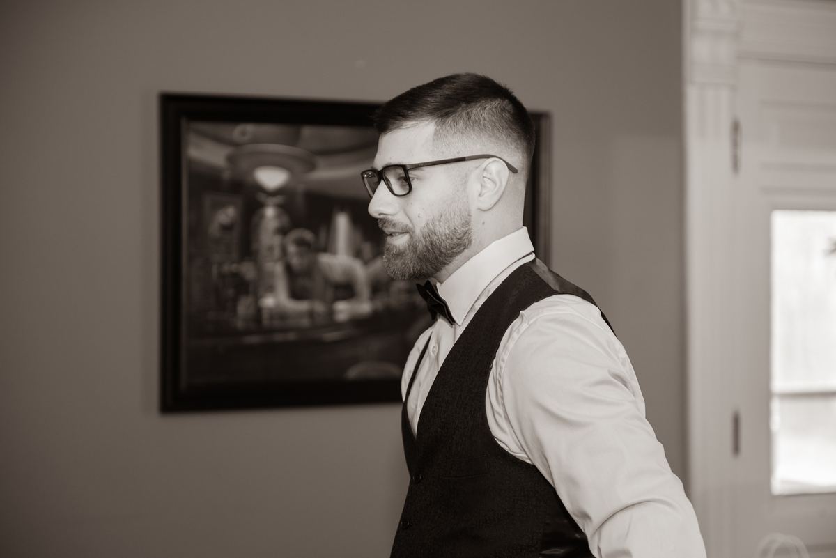 A formally dressed man smiles gently in a softly lit room with a window and framed photo behind him.