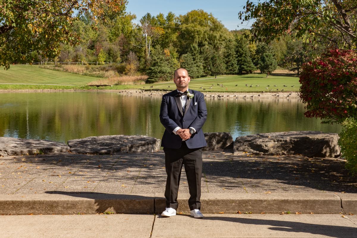 A man in wedding attire stands on a stone ledge by a pond, surrounded by autumn leaves and golf course trees.