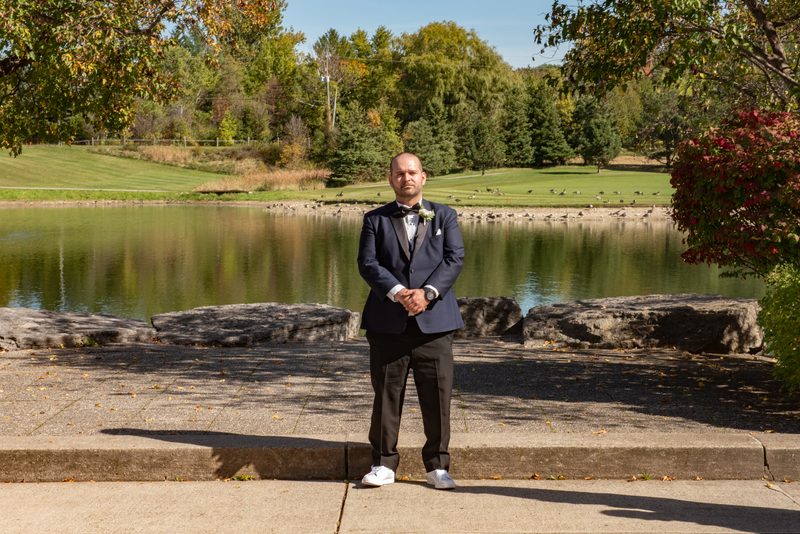 A man in wedding attire stands on a stone ledge by a pond, surrounded by autumn leaves and golf course trees.