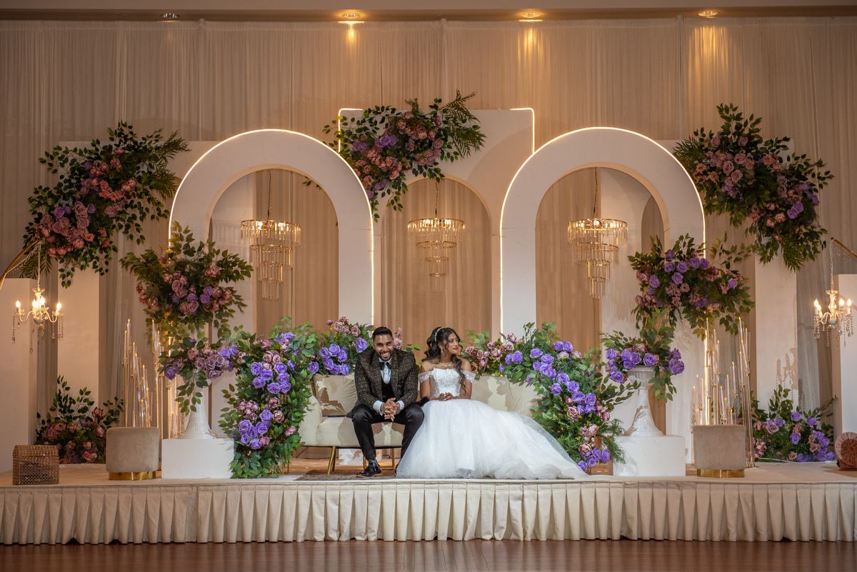 A newlywed couple sits on a decorated stage under arched backdrops with floral arrangements, illuminated by warm lights.