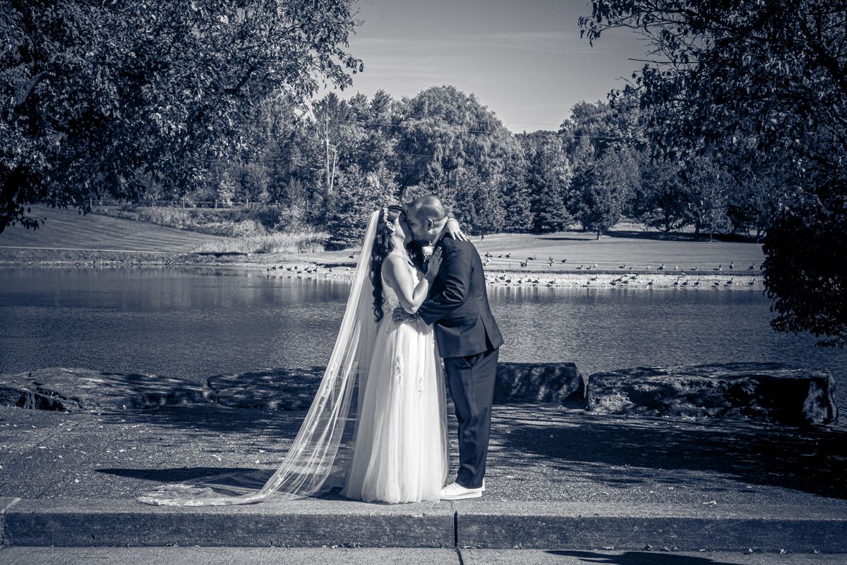 A newlywed couple in dark suit and flowing gown kiss tenderly by a calm lake, framed by trees and birds.