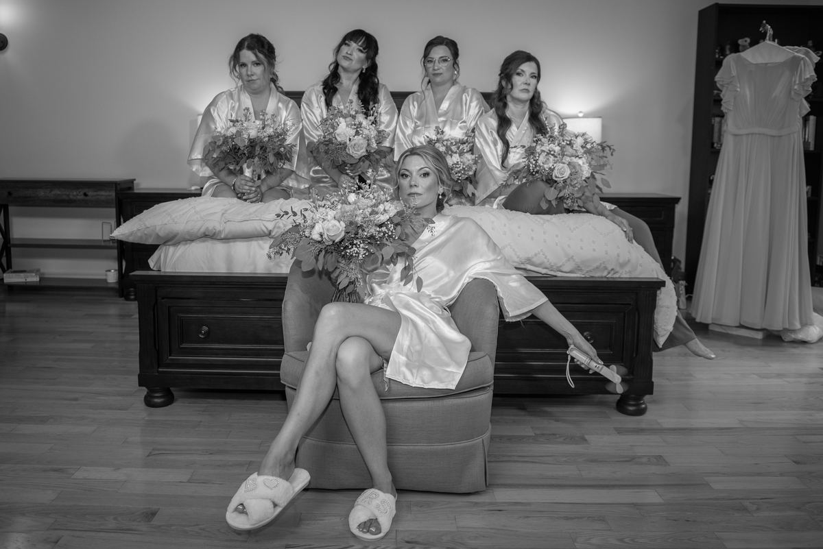 A bride sits in a chair looking at the camera while bridesmaids sit on a bed behind her holding bouquets.