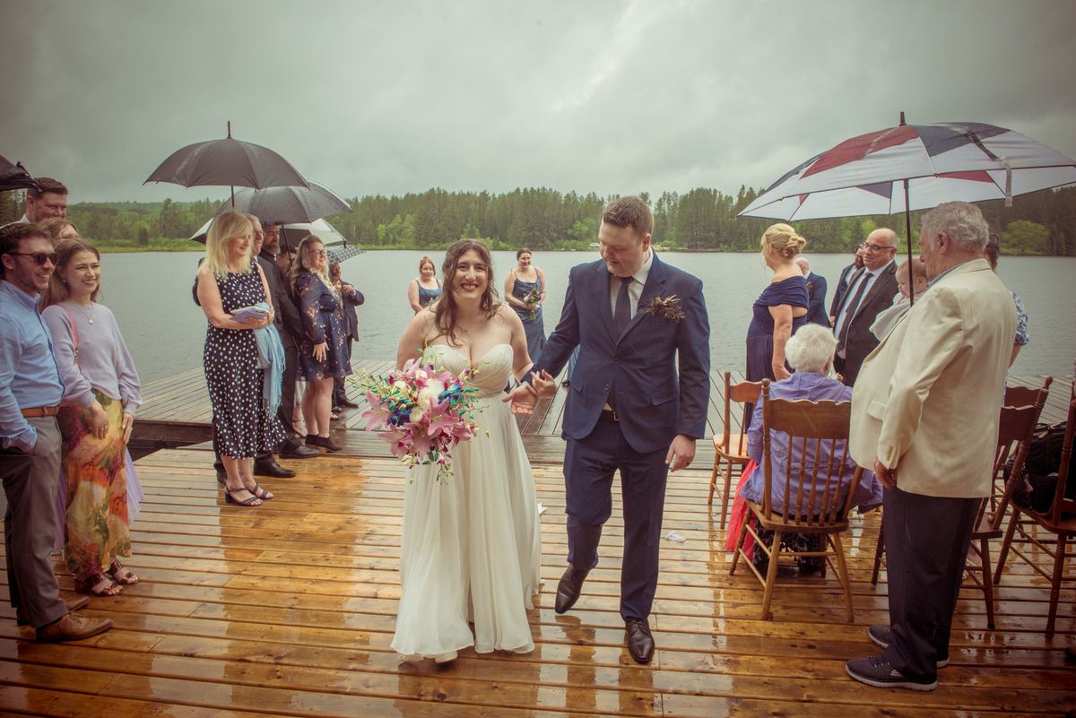 Bride and groom walk along a wet dock toward guests holding umbrellas under an overcast sky, smiling.