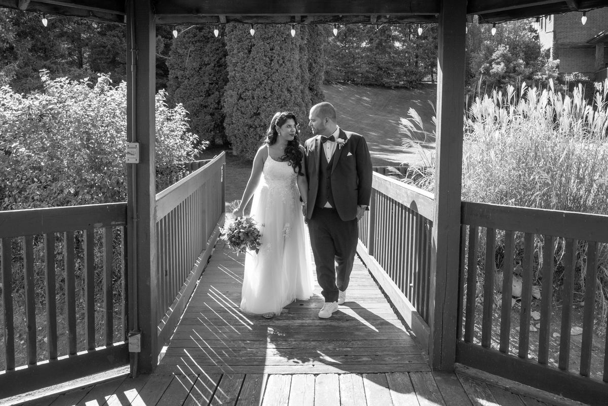 A bride and groom walk hand-in-hand on a wooden bridge in a lush garden, framed by pillars and railings under string lights.